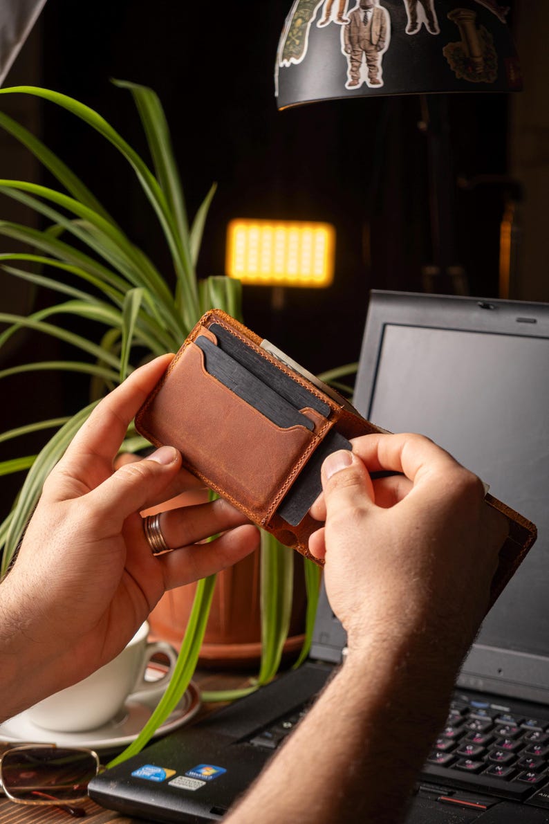 May include: A brown leather wallet with multiple card slots is being held open, revealing dark cards. The wallet is a warm, medium brown color and appears to be made of high-quality leather. The background includes a laptop and a green plant.