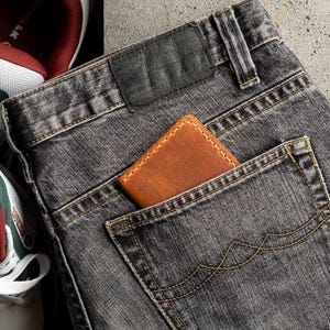 May include: Close-up of a pair of gray denim jeans with a brown leather wallet in the back pocket. The jeans have a black leather patch and are next to a pair of white and red sneakers.