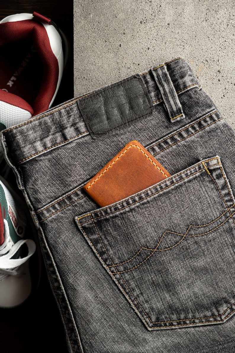 May include: Close-up of a pair of gray denim jeans with a brown leather wallet in the back pocket. The jeans have a black leather patch and are next to a pair of white and red sneakers.