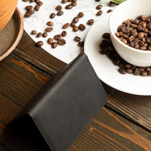 May include: A black leather wallet is open on a dark wooden surface. Coffee beans are scattered around a white bowl filled with more beans. A small terracotta pot with a plant is in the background.
