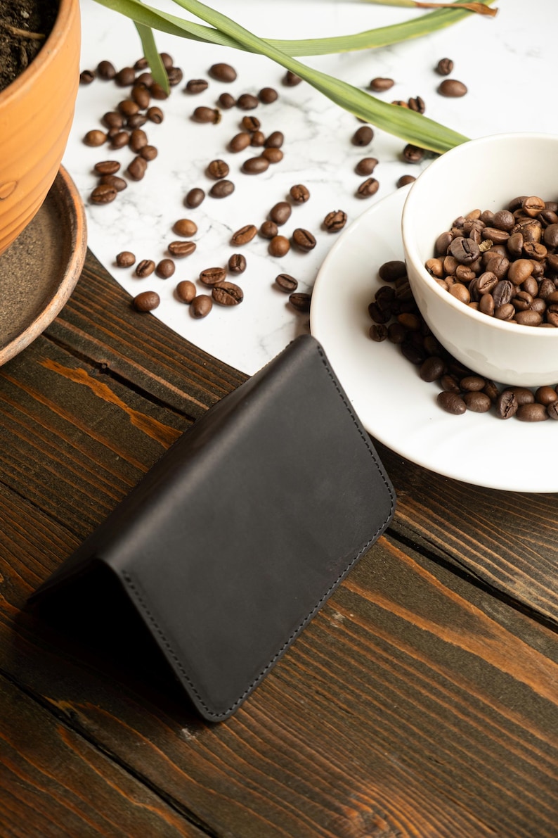 May include: A black leather wallet is open on a dark wooden surface. Coffee beans are scattered around a white bowl filled with more beans. A small terracotta pot with a plant is in the background.