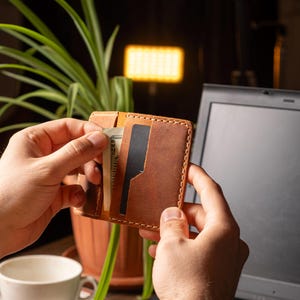 May include: A brown leather wallet with visible stitching, held open to reveal a cash and a black card. The wallet is in front of a laptop and a potted plant. The wallet is a bi-fold style.