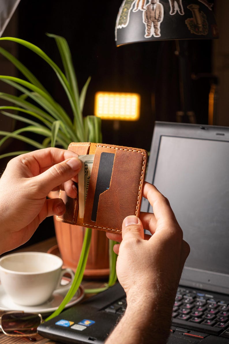 May include: A brown leather wallet with visible stitching, held open to reveal a cash and a black card. The wallet is in front of a laptop and a potted plant. The wallet is a bi-fold style.