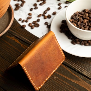 May include: A brown leather wallet with white stitching is open on a dark wooden surface. Coffee beans are scattered around a white bowl filled with more beans. A terracotta pot with a plant is in the background.