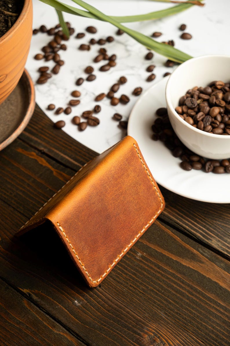 May include: A brown leather wallet with white stitching is open on a dark wooden surface. Coffee beans are scattered around a white bowl filled with more beans. A terracotta pot with a plant is in the background.