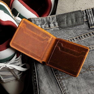 May include: Open, brown leather wallet with multiple card slots, resting on a pair of gray denim jeans. The wallet is displayed open, showcasing its interior design. A pair of sneakers with white, green, and burgundy accents are in the background.