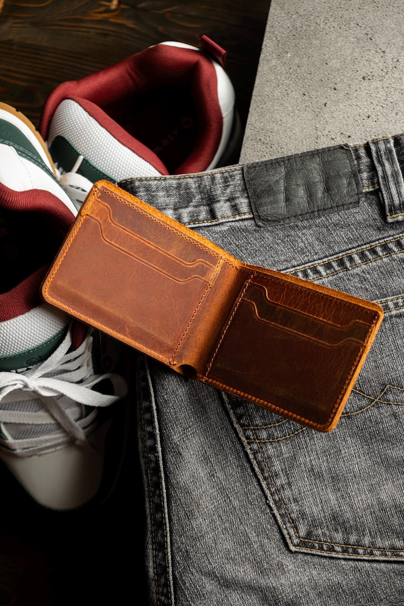 May include: Open, brown leather wallet with multiple card slots, resting on a pair of gray denim jeans. The wallet is displayed open, showcasing its interior design. A pair of sneakers with white, green, and burgundy accents are in the background.