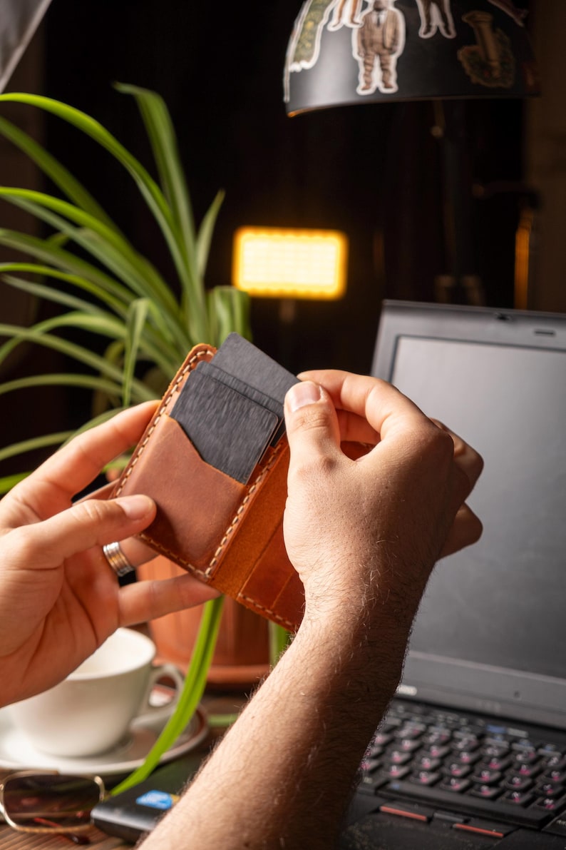 May include: A brown leather cardholder with a lighter brown pocket is held open, revealing several dark gray cards. The cardholder is stitched with a light-colored thread. The background includes a laptop, a cup, and a plant.