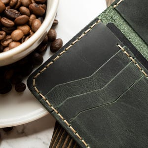 May include: Dark green leather wallet with cream-colored stitching. The wallet has multiple card slots and a textured surface. A white coffee cup filled with coffee beans is in the background.