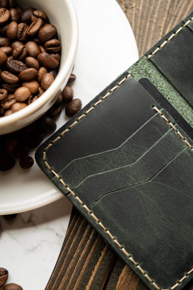 May include: Dark green leather wallet with cream-colored stitching. The wallet has multiple card slots and a textured surface. A white coffee cup filled with coffee beans is in the background.