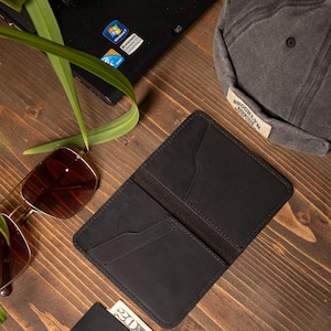 May include: Open black leather wallet with card slots, next to a black cap with "BROOKLYN" text, sunglasses, a laptop, and a Visa card. The items are arranged on a wooden surface.