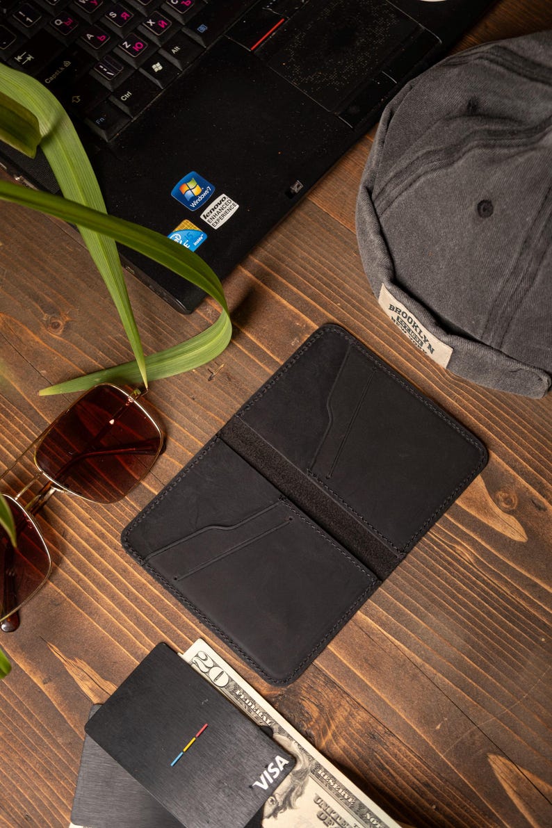May include: Open black leather wallet with card slots, next to a black cap with "BROOKLYN" text, sunglasses, a laptop, and a Visa card. The items are arranged on a wooden surface.