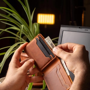 May include: A brown leather wallet is open, revealing card slots and a bill compartment. A twenty-dollar bill is being inserted into the wallet. The wallet is on a laptop keyboard, with a plant and a lamp in the background.