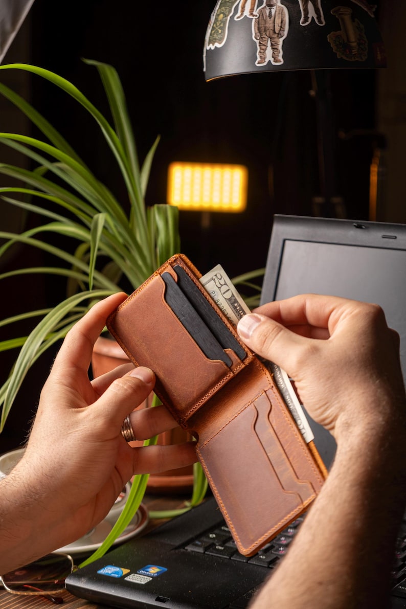 May include: A brown leather wallet is open, revealing card slots and a bill compartment. A twenty-dollar bill is being inserted into the wallet. The wallet is on a laptop keyboard, with a plant and a lamp in the background.
