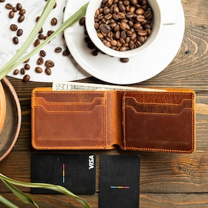 May include: Open brown leather wallet with multiple card slots and a bill inside. Two black cards with colorful stripes and the word "VISA" are next to the wallet. A white cup filled with coffee beans sits on a white plate. Coffee beans and greenery are scattered around.