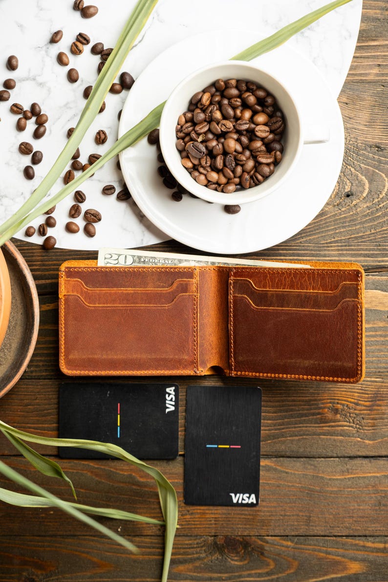 May include: Open brown leather wallet with multiple card slots and a bill inside. Two black cards with colorful stripes and the word "VISA" are next to the wallet. A white cup filled with coffee beans sits on a white plate. Coffee beans and greenery are scattered around.