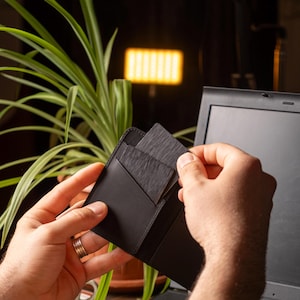 May include: A black leather wallet with several dark gray cards being removed. The wallet is held in front of a laptop, with a plant and a lamp in the background. The wallet appears to be new and unused.