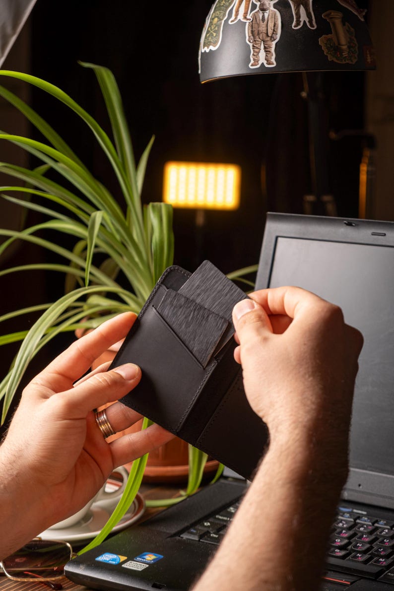 May include: A black leather wallet with several dark gray cards being removed. The wallet is held in front of a laptop, with a plant and a lamp in the background. The wallet appears to be new and unused.