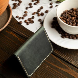 May include: Dark green leather wallet with white stitching, resting on a wooden surface. A white cup and saucer filled with coffee beans are in the background, along with scattered beans. The wallet is a minimalist design.