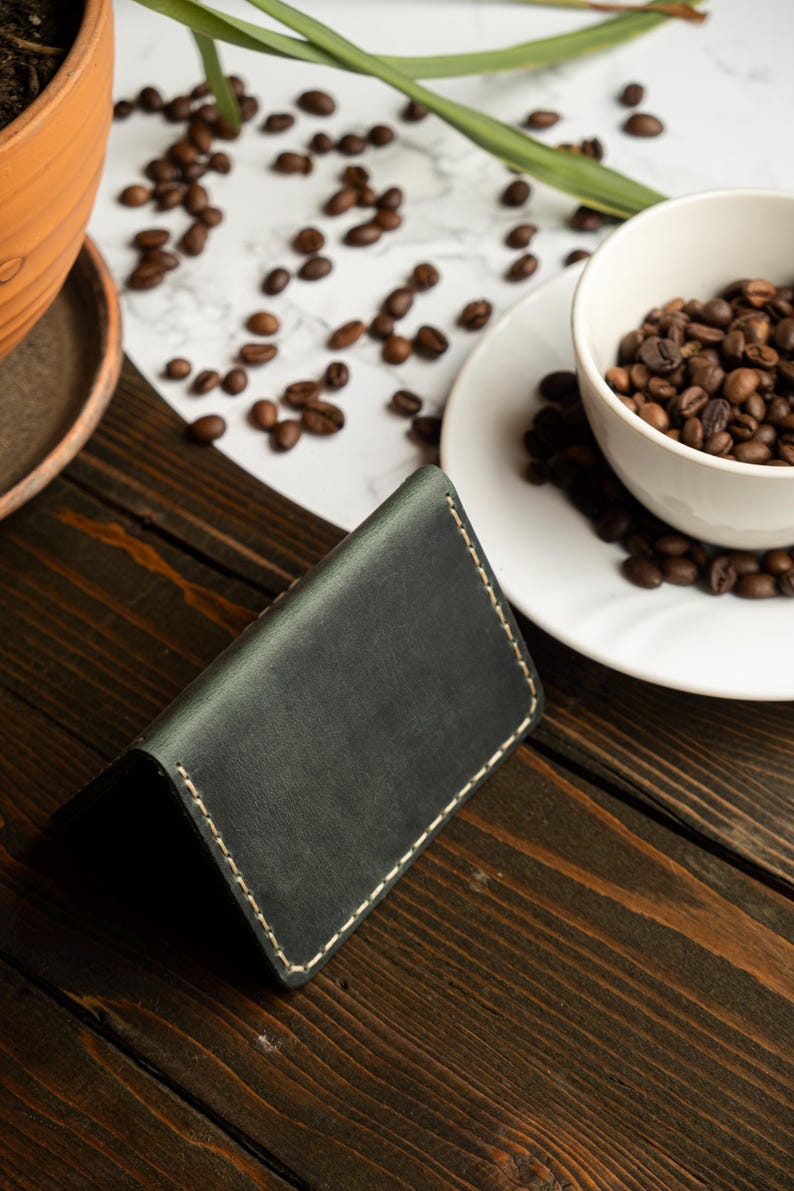 May include: Dark green leather wallet with white stitching, resting on a wooden surface. A white cup and saucer filled with coffee beans are in the background, along with scattered beans. The wallet is a minimalist design.