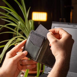 May include: A black leather wallet with a contrasting light-colored stitch detail. The wallet is open, revealing several dark gray cards being inserted into a pocket. The background includes a laptop and a green plant.