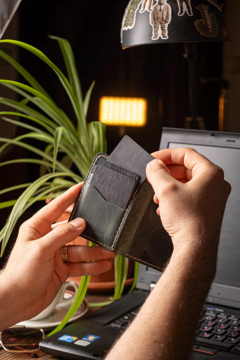 May include: A black leather wallet with a contrasting light-colored stitch detail. The wallet is open, revealing several dark gray cards being inserted into a pocket. The background includes a laptop and a green plant.