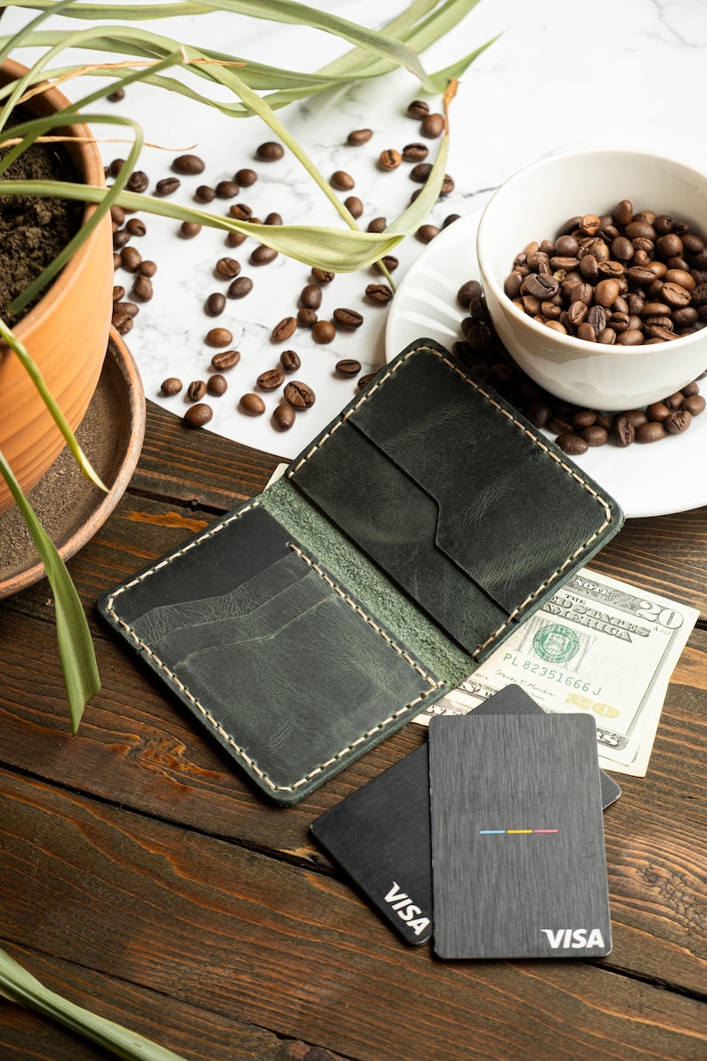 May include: Open, dark green leather wallet with cream stitching, lying open on a wooden surface. Two black credit cards with the Visa logo and a colorful stripe design are next to the wallet. A cup of coffee beans and a US dollar bill are also visible.