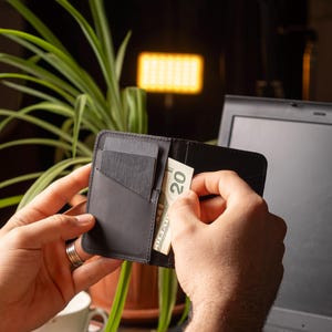 May include: Black leather wallet with card slots and a bill compartment. The wallet is open, revealing a twenty-dollar bill. The background includes a laptop, a plant, and a cup of coffee.