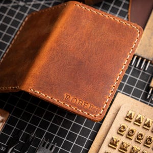 May include: A handcrafted brown leather wallet with the name "ROBERT" embossed on the front. The wallet features white stitching and is surrounded by leatherworking tools, including stamps and thread spools.