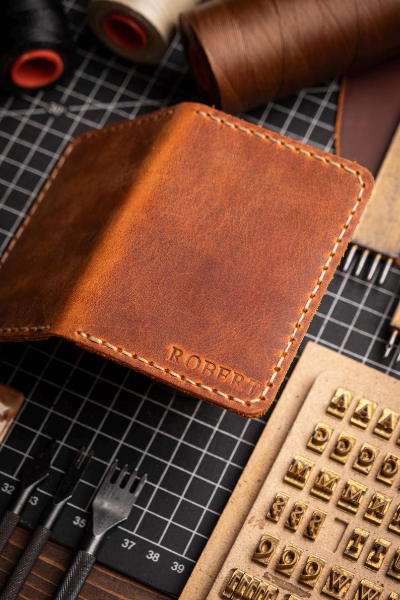 May include: A handcrafted brown leather wallet with the name "ROBERT" embossed on the front. The wallet features white stitching and is surrounded by leatherworking tools, including stamps and thread spools.