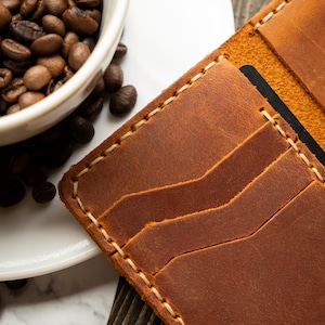 May include: A close-up of a brown leather wallet with multiple card slots, stitched with cream-colored thread. A white ceramic cup filled with coffee beans sits next to the wallet on a wooden surface.