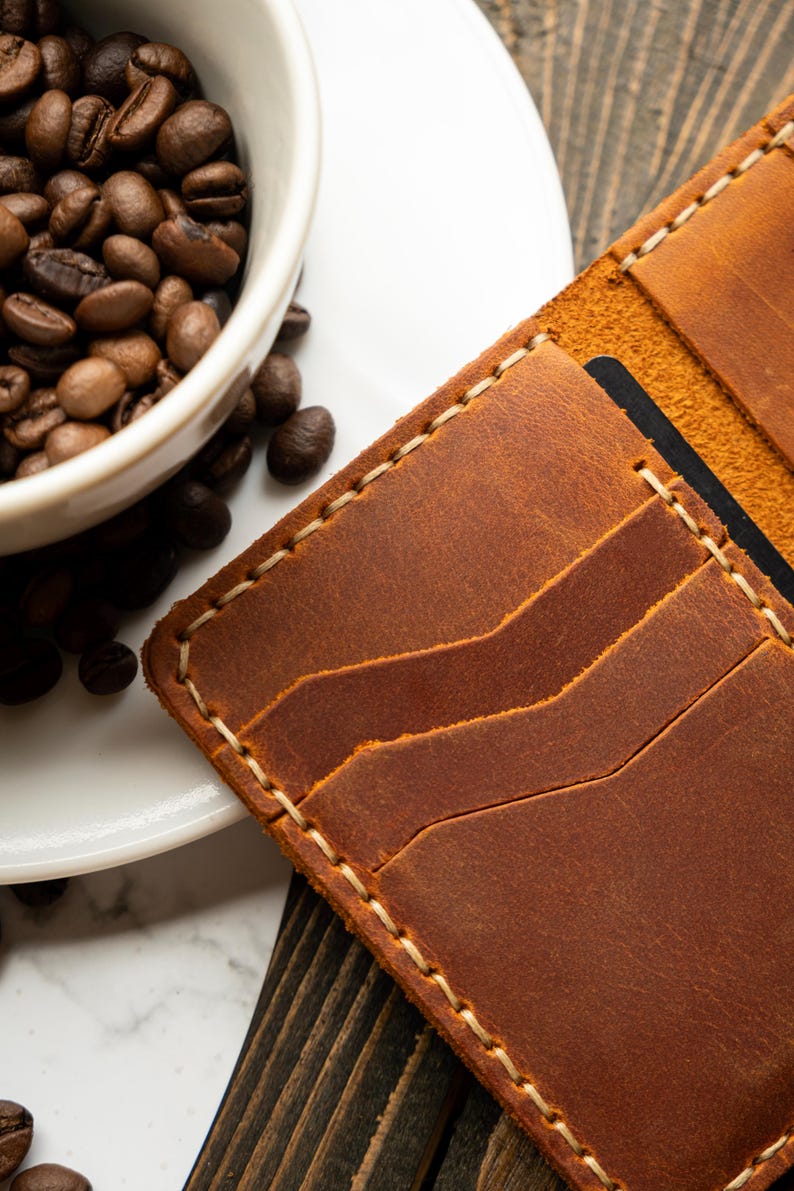 May include: A close-up of a brown leather wallet with multiple card slots, stitched with cream-colored thread. A white ceramic cup filled with coffee beans sits next to the wallet on a wooden surface.