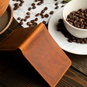 May include: A brown leather wallet with a textured surface, open to reveal the interior. The wallet is on a dark wooden surface, with coffee beans scattered around a white cup and saucer. A terracotta pot with a plant is in the background.