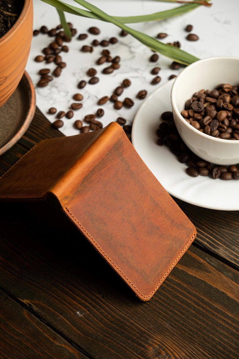 May include: A brown leather wallet with a textured surface, open to reveal the interior. The wallet is on a dark wooden surface, with coffee beans scattered around a white cup and saucer. A terracotta pot with a plant is in the background.