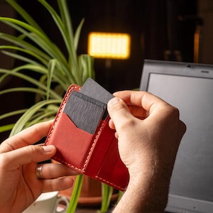 May include: A red leather wallet with white stitching, holding several dark gray cards. The wallet is open, and cards are being inserted or removed. The background includes a laptop, a plant, and a cup.
