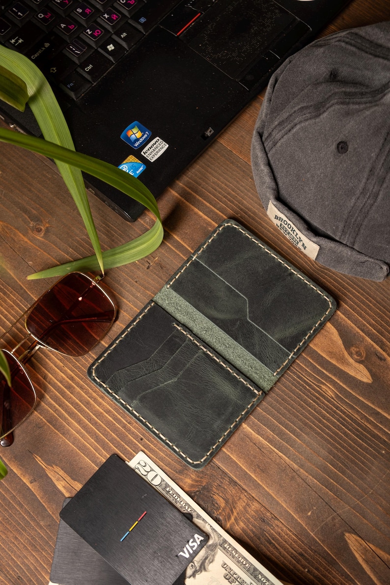 May include: Open, dark green leather wallet with white stitching, next to a black credit card and a twenty-dollar bill. A pair of sunglasses and a gray cap are also visible.