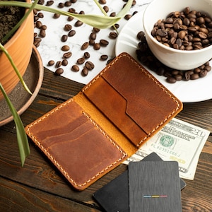 May include: Open, brown leather wallet with multiple card slots, set on a wooden surface. A twenty-dollar bill and two Visa cards are next to the wallet. A cup of coffee beans and a potted plant are in the background.