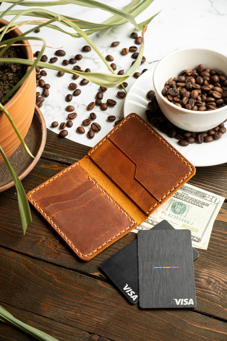May include: Open, brown leather wallet with multiple card slots, set on a wooden surface. A twenty-dollar bill and two Visa cards are next to the wallet. A cup of coffee beans and a potted plant are in the background.
