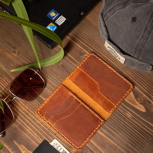 May include: Open brown leather wallet with multiple card slots, stitched with white thread. A pair of sunglasses, a gray cap with "BROOKLYN" text, a laptop, a credit card, and a twenty-dollar bill are also visible.