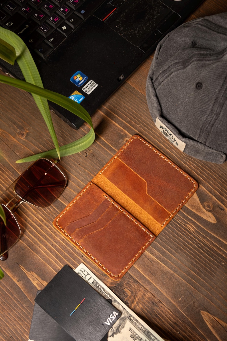 May include: Open brown leather wallet with multiple card slots, stitched with white thread. A pair of sunglasses, a gray cap with "BROOKLYN" text, a laptop, a credit card, and a twenty-dollar bill are also visible.