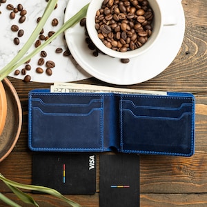 May include: Open navy blue leather wallet with multiple card slots and a cash compartment. Two black credit cards with a colorful stripe design are next to the wallet. A cup of coffee beans sits on a white plate.
