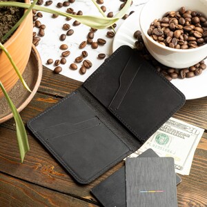 May include: Open black leather wallet with card slots, next to two gray cards with the Visa logo and a colorful stripe design. A twenty-dollar bill is visible. The scene includes coffee beans and a small plant.