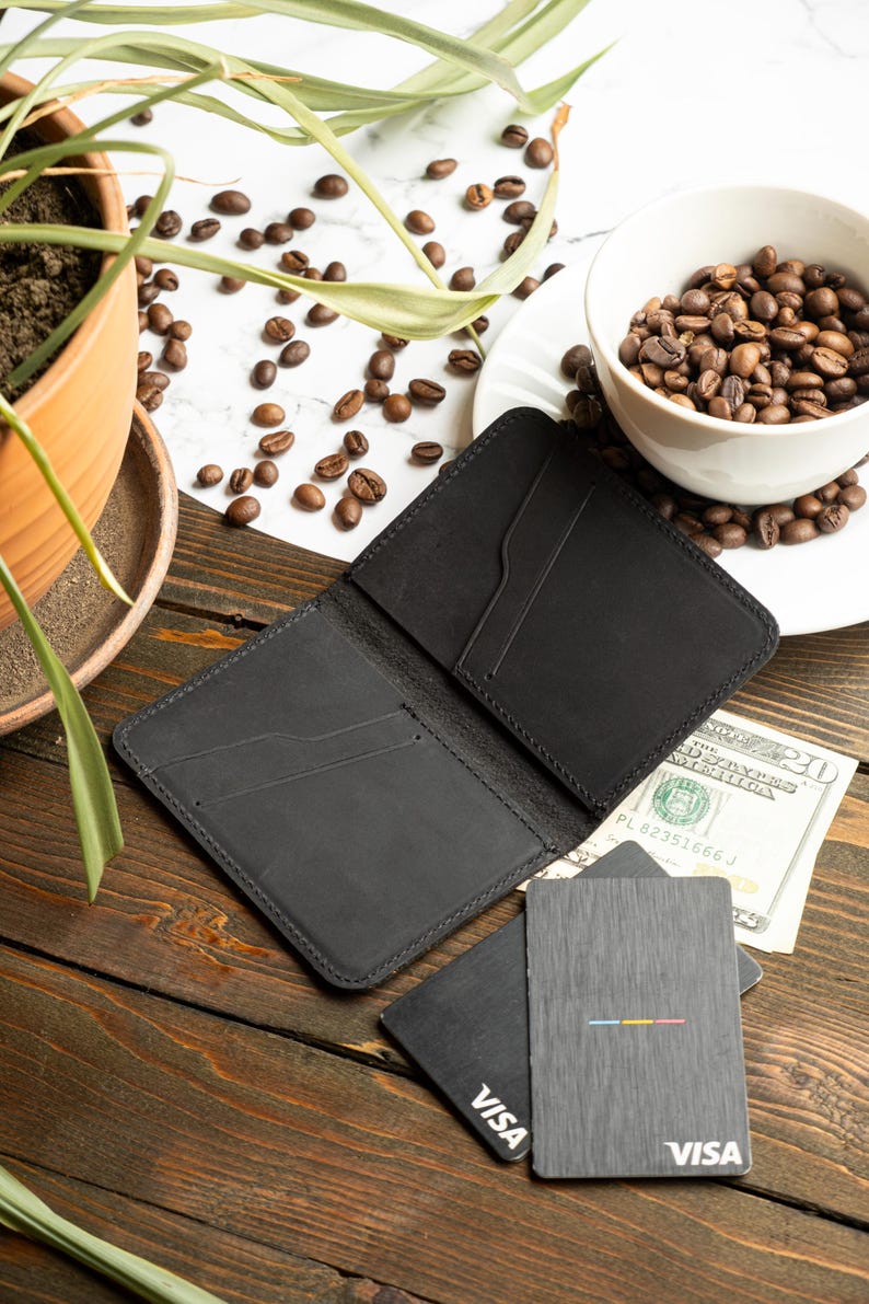 May include: Open black leather wallet with card slots, next to two gray cards with the Visa logo and a colorful stripe design. A twenty-dollar bill is visible. The scene includes coffee beans and a small plant.