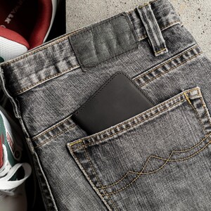 May include: Close-up of gray denim jeans with a black leather wallet in the back pocket. The jeans have yellow stitching and a leather patch. A pair of sneakers with red and white accents are visible in the background.