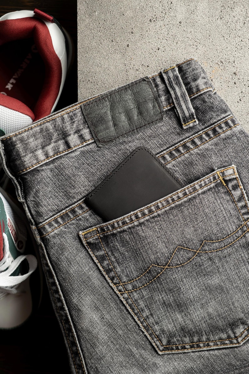 May include: Close-up of gray denim jeans with a black leather wallet in the back pocket. The jeans have yellow stitching and a leather patch. A pair of sneakers with red and white accents are visible in the background.