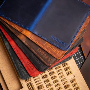 May include: A stack of personalized leather wallets in various colors, including blue, brown, red, and black. Each wallet is embossed with the name "ROBERT" in a matching color. A wooden block with metal letter stamps is in the foreground.