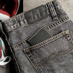 May include: Close-up of a pair of gray denim jeans with a black leather patch and a wallet in the back pocket. The jeans have yellow stitching and are next to a pair of sneakers.