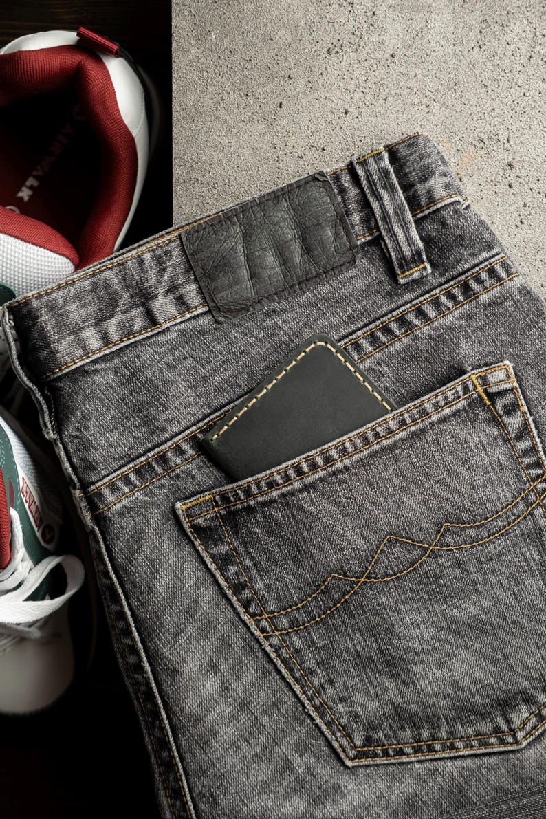 May include: Close-up of a pair of gray denim jeans with a black leather patch and a wallet in the back pocket. The jeans have yellow stitching and are next to a pair of sneakers.