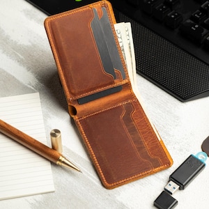May include: Open brown leather wallet with multiple card slots and cash. The wallet is on a light-colored surface with a pen, notepad, and a USB drive. The wallet is filled with cash and cards.