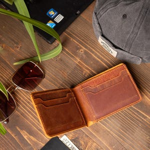 May include: Open, brown leather wallet with multiple card slots, resting on a wooden surface. A pair of sunglasses, a gray cap, a laptop, and a Visa card are also visible. The wallet is empty.